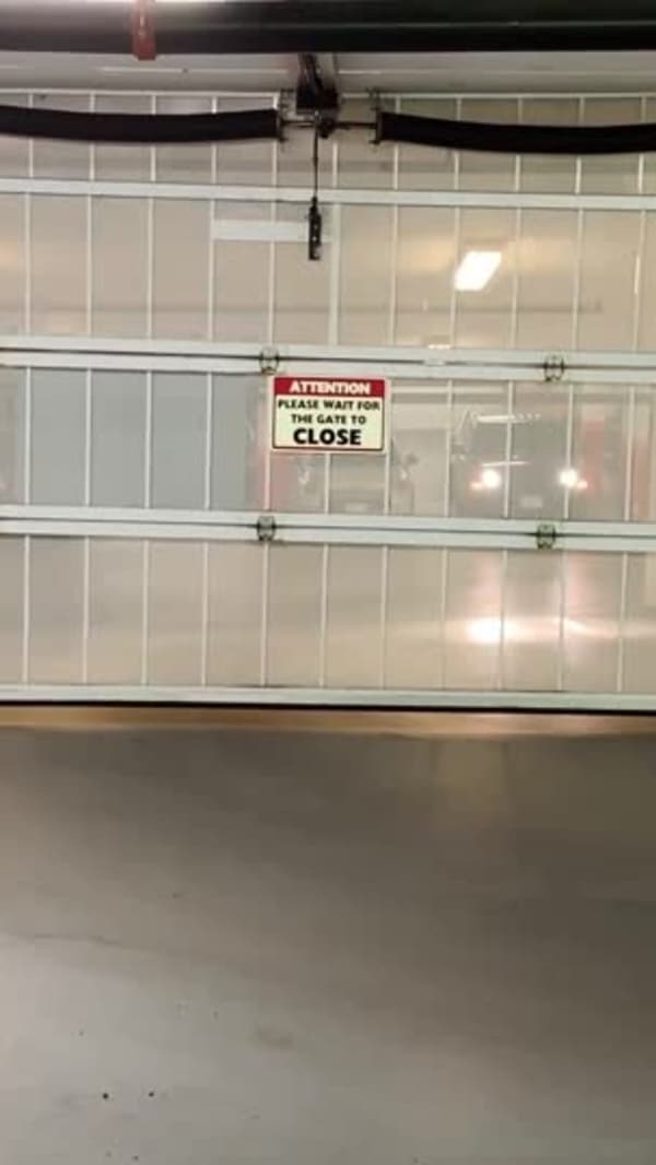 Open parkade security gate seen from inside the garage, headlights of a waiting car visible through the slats and bare concrete floor in the foreground