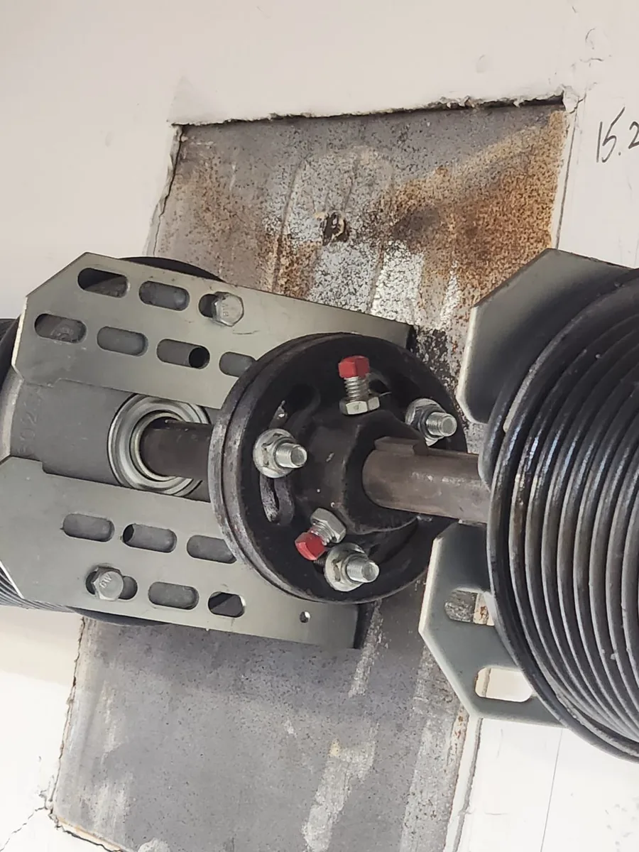 Torsion shaft end on a commercial door: end bearing plate, 4-inch cable drum with red set screws, and the torsion spring beside it